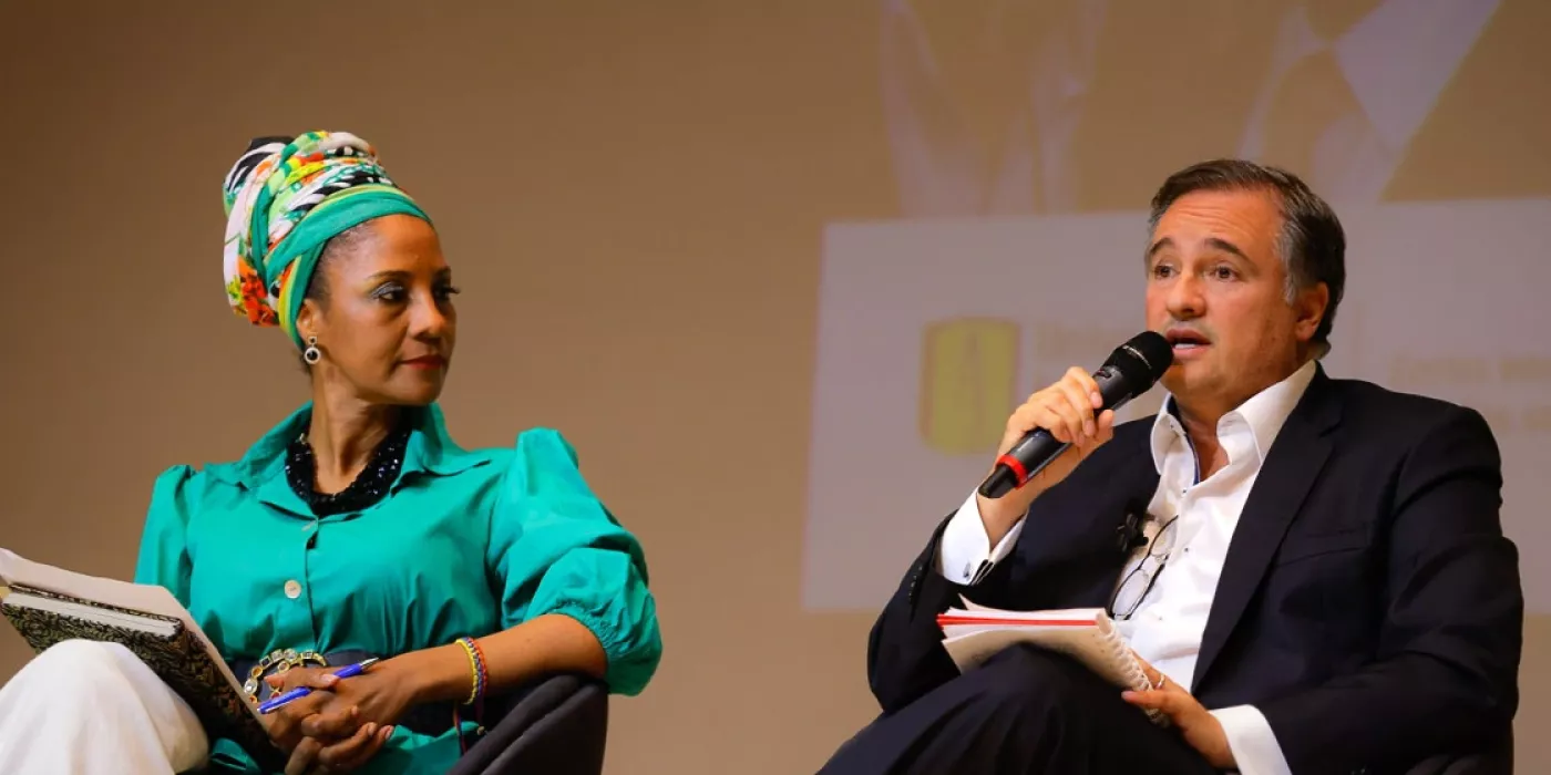 Hombre habla por micrófono. Una mujer lo mira.