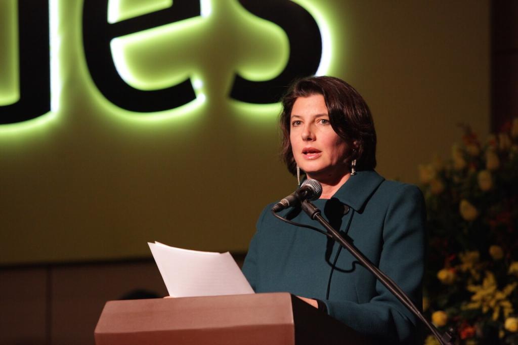 una mujer de abrigo verde oscuro da un discurso frente a un atril