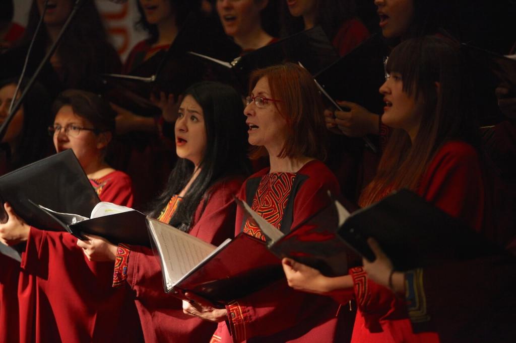 unas mujeres de un coro vestidas con batas vinotinto interpretan un himno