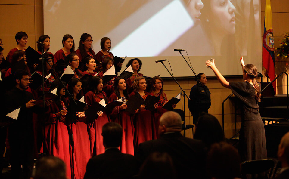 Una mujer de cabello largo vestida de negro dirige  un coro de mujeres con vestido vinotinto