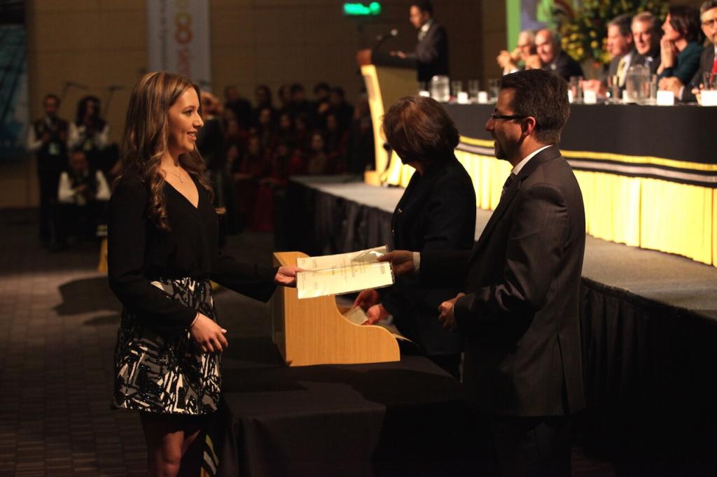 una joven de vestido negro con gris recibe diploma de grado en ceremonia en un auditorio