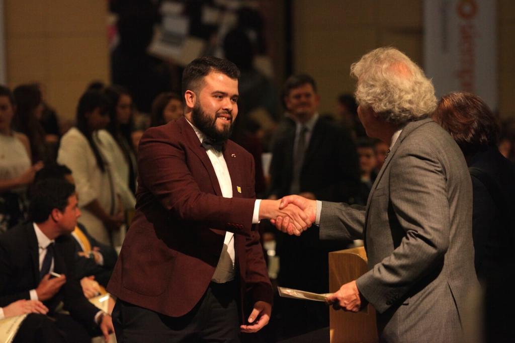 un joven de saco vinotinto recibe diploma y da la mano a un hombre mayor