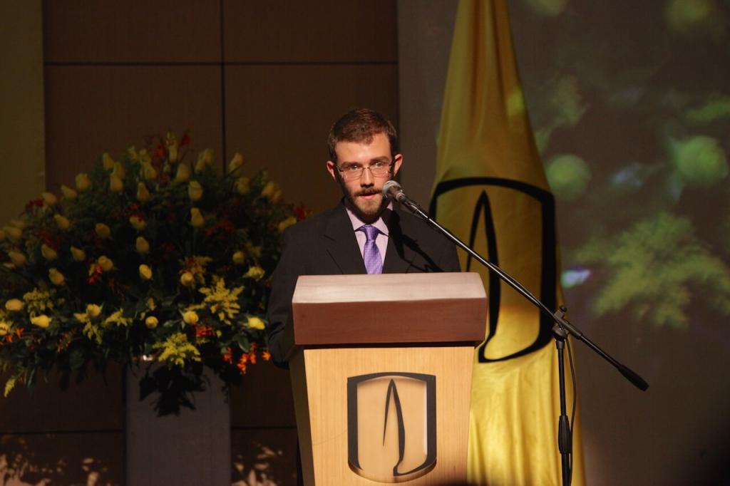 un joven estudiante con gafas y barba habla frente a un micrófono en una ceremonia de grados