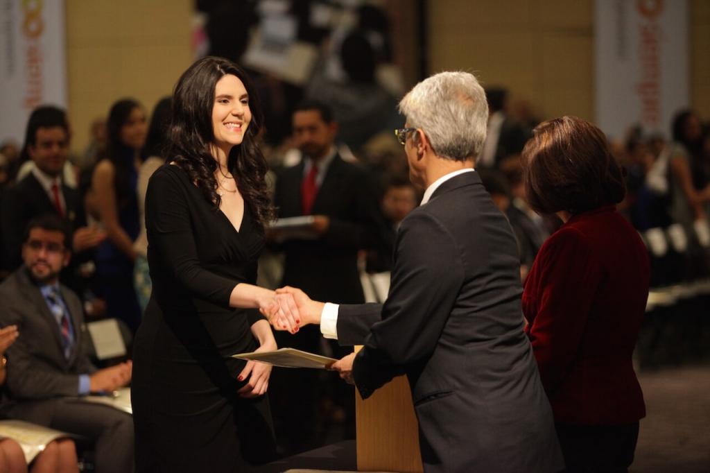 Mujer alta de cabello negro y vestido largo estrecha la mano de un hombre mayor al recibir diploma