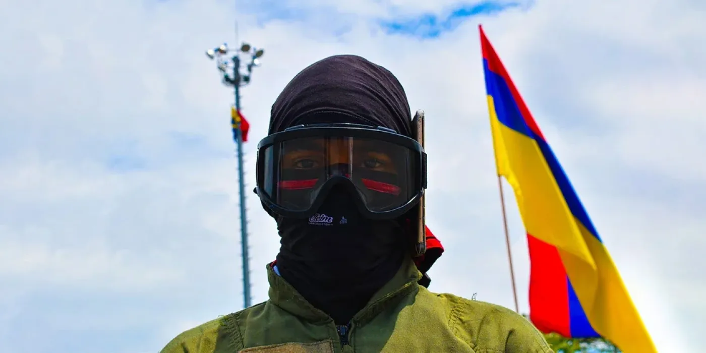"Hombre con capucha en primer plano. Atrás la bandera de Colombia