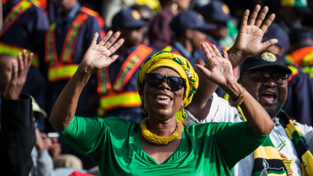 Mujer sudafricana con vestido verde y pañoleta amarilla sonríe y alza sus manos. 